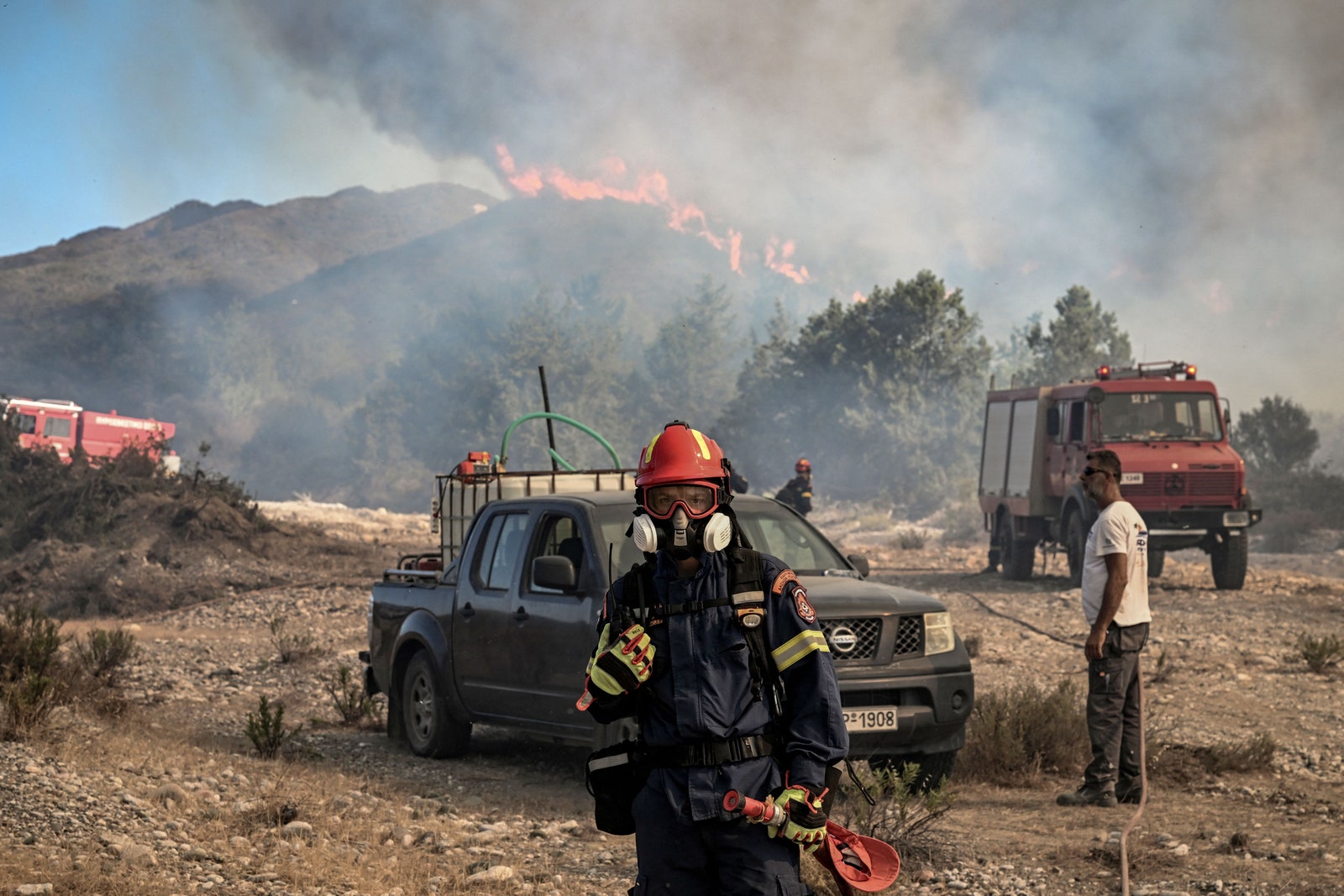 マスクを着けた消防士が、バティ村の近くの丘陵地帯の森林が山火事で焼け落ちる様子を見守っている。